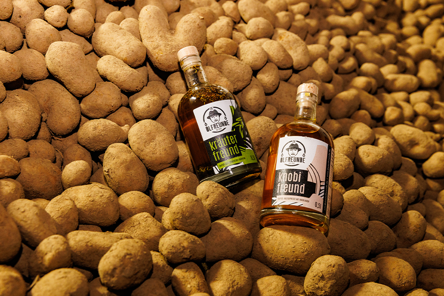 2 Ölflachen mit grünem und rosa Etikett liegen in einem Stapel von Kartoffeln.