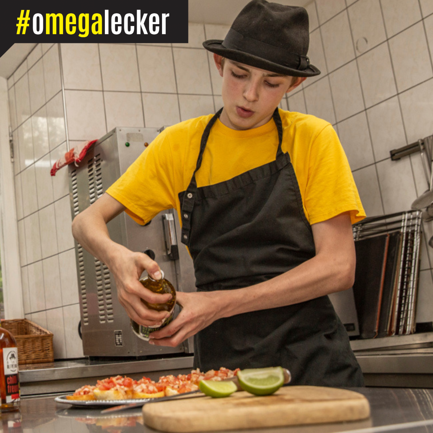 Eine Person in einer Küche, die ein gelbes T-Shirt, eine schwarze Schürze und einen schwarzen Hut trägt. Die Person hält eine Flasche und gießt deren Inhalt über einen Teller, der mit Bruschetta aus gewürfelten Tomaten belegt ist. Im Vordergrund liegt ein Schneidebrett mit zwei Limettenkeilen