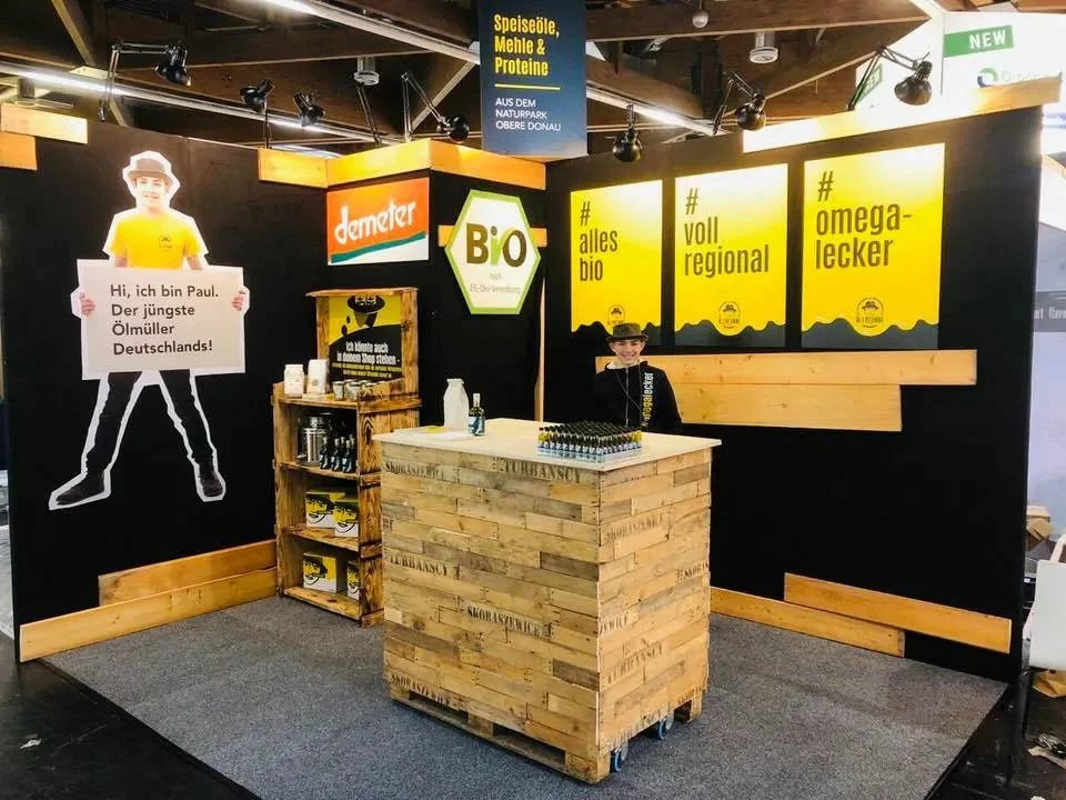 Ein Messestand, der regionale und biologische Speiseöle, Mehle & Proteine aus dem Naturpark Obere Donau bewirbt. Im Vordergrund zeigt ein großer Aufsteller die Aufschrift 'Hi, ich bin Paul. Der jüngste Ölmüller Deutschlands!'. An der Wand befinden sich drei Poster mit Hashtags. Über dem Stand prangt ein Schild mit der Produktüberschrift, ergänzt durch Demeter- und Bio-Logos.