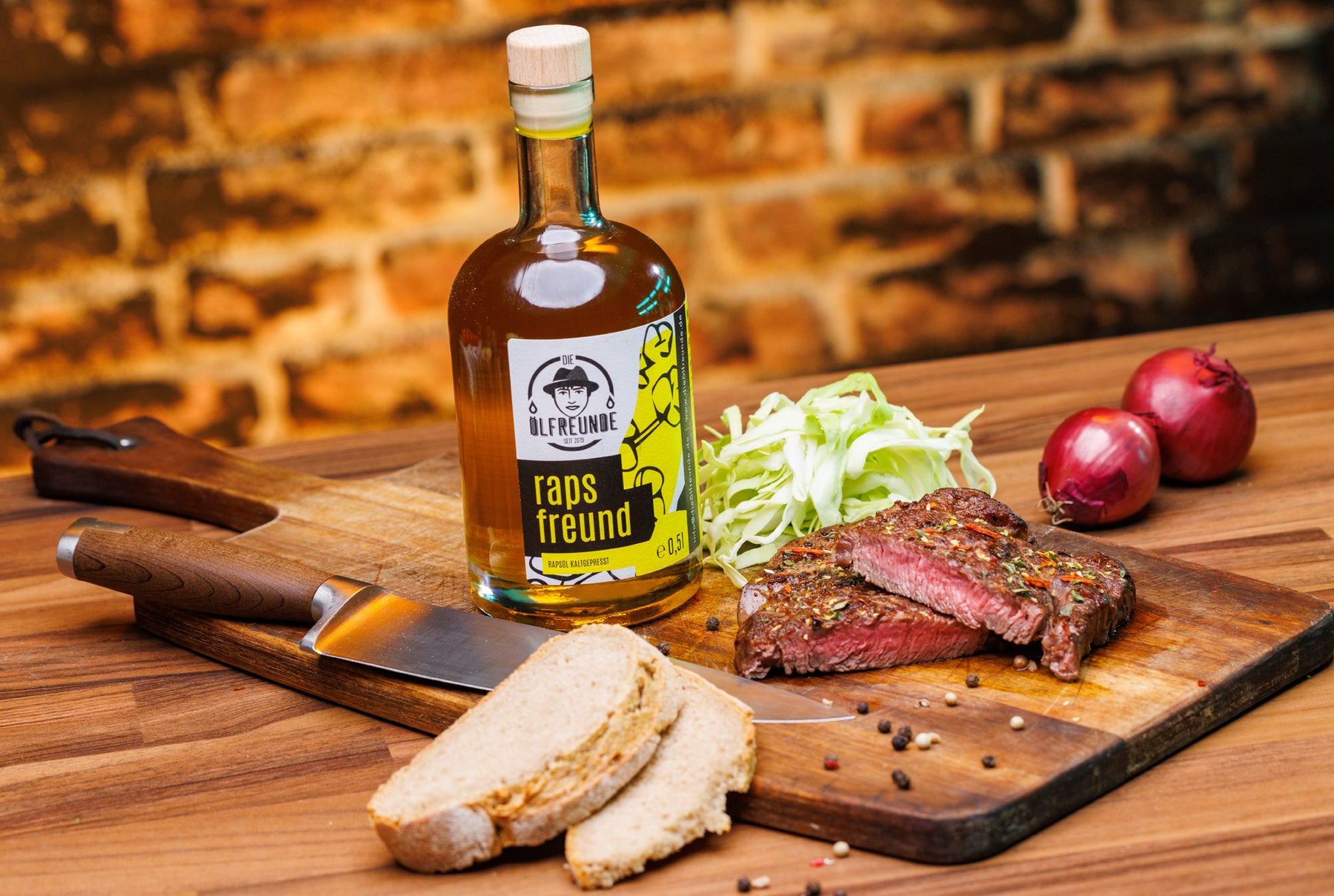 Eine Flasche mit 500ml Rapsöl der Marke Die Ölfreunde, auf einem Holzbrett, neben einem Stück Steak, Salat, Brot, Zwiebeln und einem Messer.