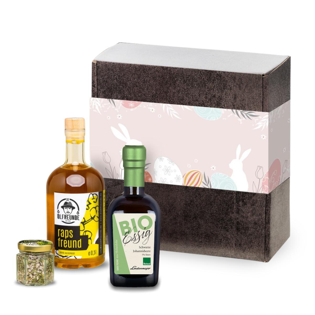 Schwarze Johannisbeere-Essig der Marke Lindenmeyer, Kräutersalz und Rapsöl der Marke Die Ölfreunde, vor einer Oster-Geschenkeverpackung.
