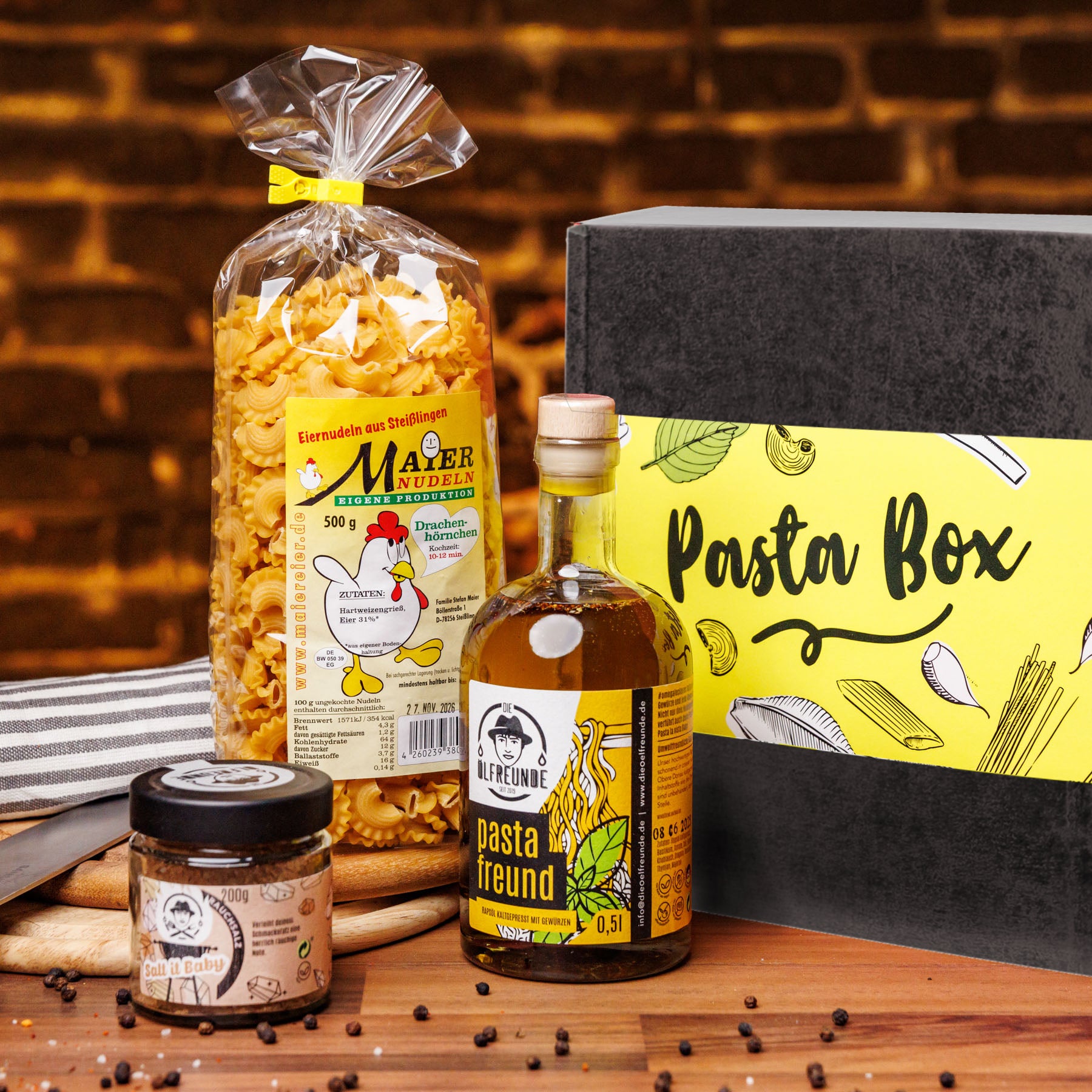 Zusammenstellung von Produkten mit Nudeln von Marke Maier Nudeln, Pasta-Öl und Rauchsalz von Marke die Ölfreunde, neben einer Pasta-Geschenkebox, auf einem Holzbrett.