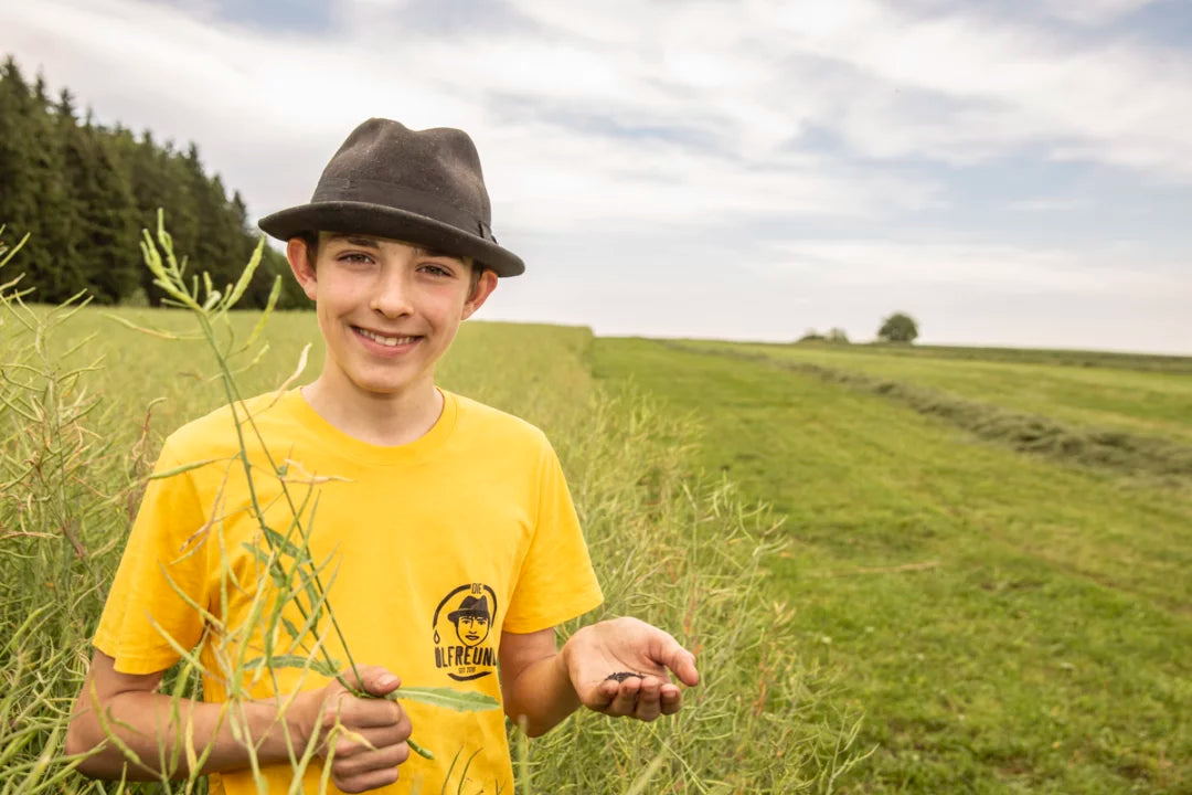 Eine Person mit schwarzem Hut und gelben T-Shirt, die auf einem Rapsfeld steht. In der einen Hand hält sie unreifen Raps und in der anderen die Rapssamen.