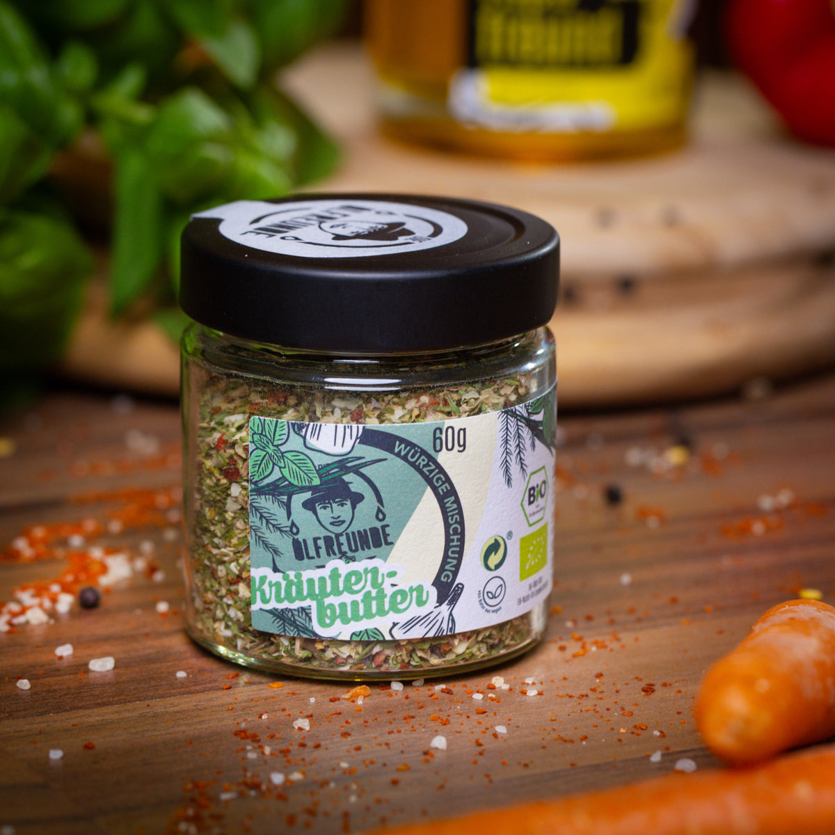 Glas mit 60 g Bio-Kräuterbutter-Gewürzmischung von der Marke Die Ölfreunde, umgeben von Gewürzen und frischen Zutaten auf einem Holzbrett.