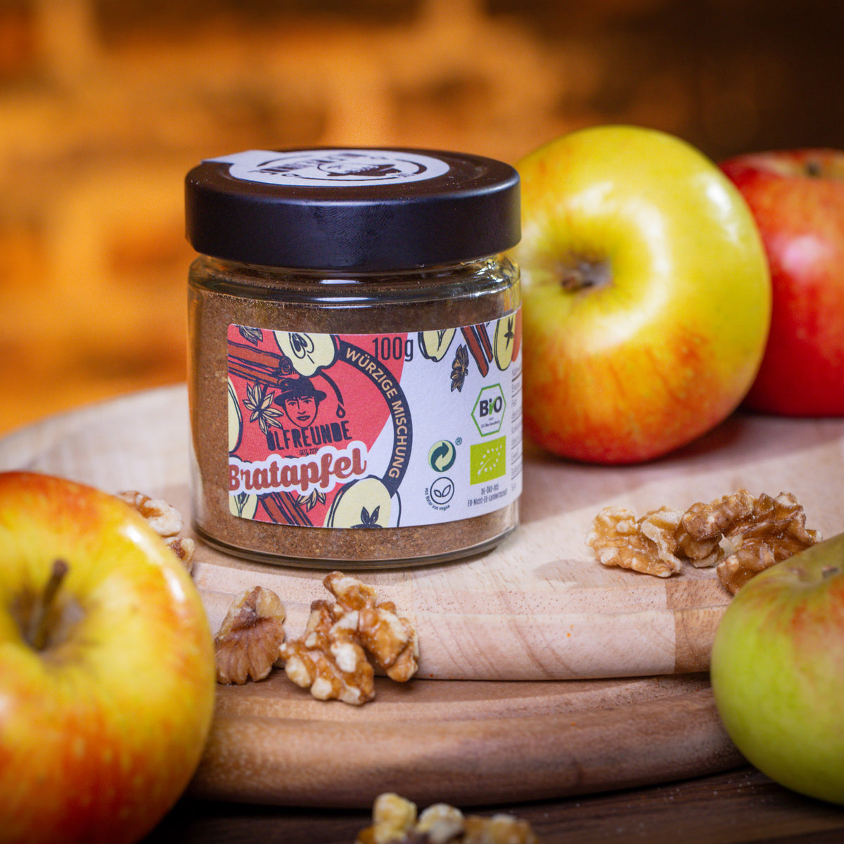 Glas mit 100 g Gewürzmischung ‚Bratapfel‘ von Die Ölfreunde, illustriertes Etikett mit Äpfeln und Gewürzen, umgeben von frischen Äpfeln und Walnüssen auf einem Holzbrett.