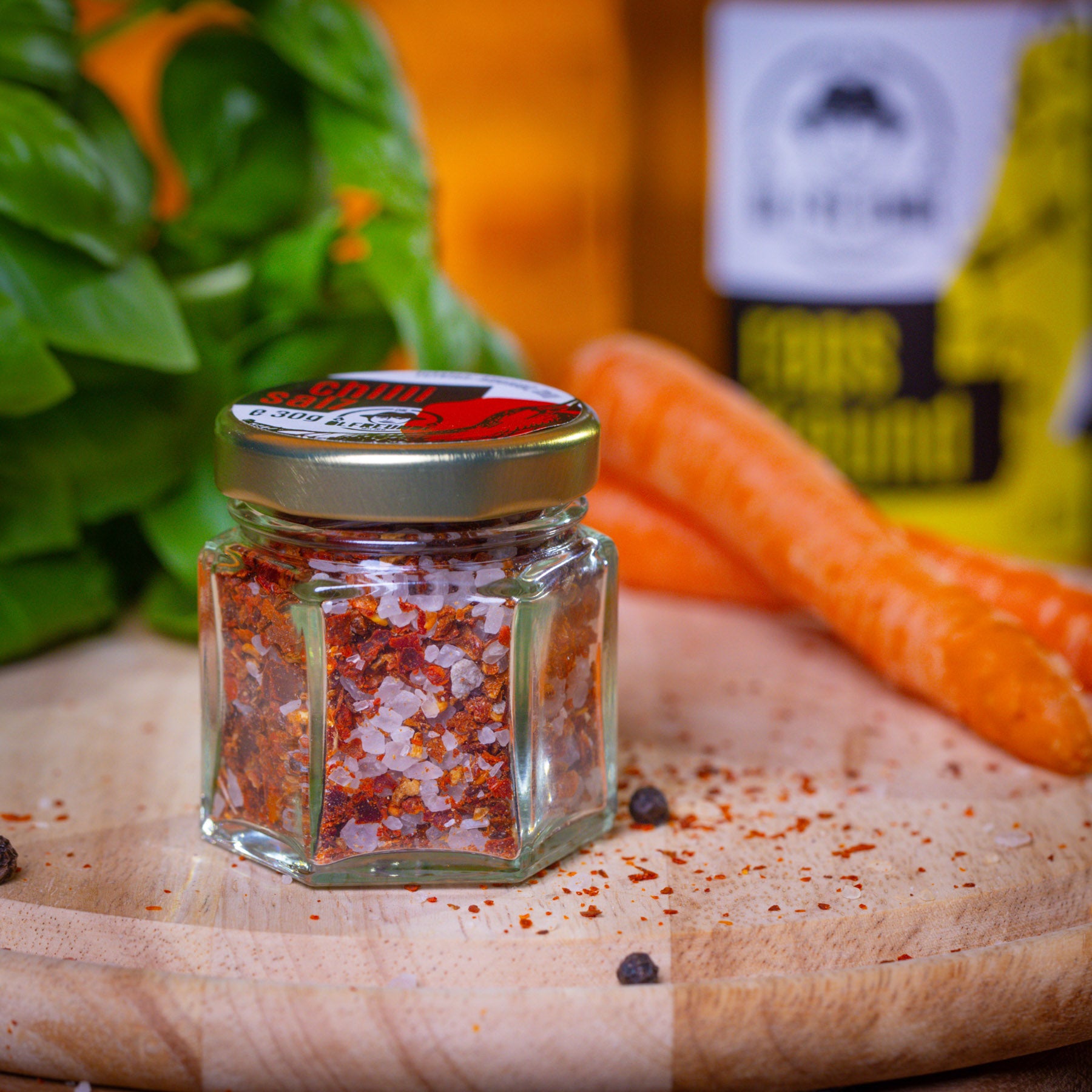 30g Chilisalz im Glas mit rotem Etikett der Marke Die Ölfeunde auf einem Holzbrett, im Hintergrund frischer Basilikum und Karotten.