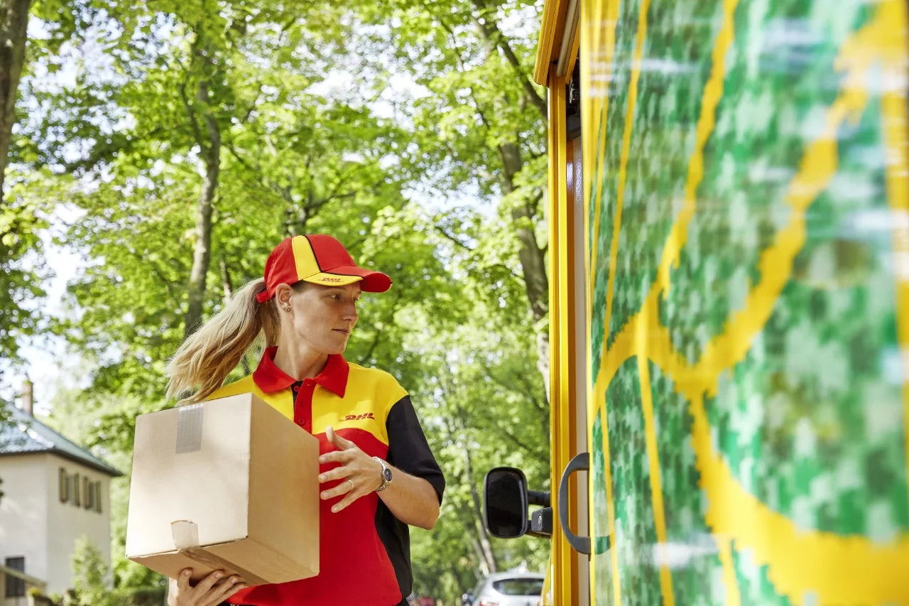 Abgebildet ist eine Person in einer rot-gelben DHL-Uniform mit sichtbarem DHL-Logo auf Shirt und Mütze. Die Person hält einen Karton in der Hand und steht neben einem gelben Lieferfahrzeug mit grün-gelben Akzentmustern.