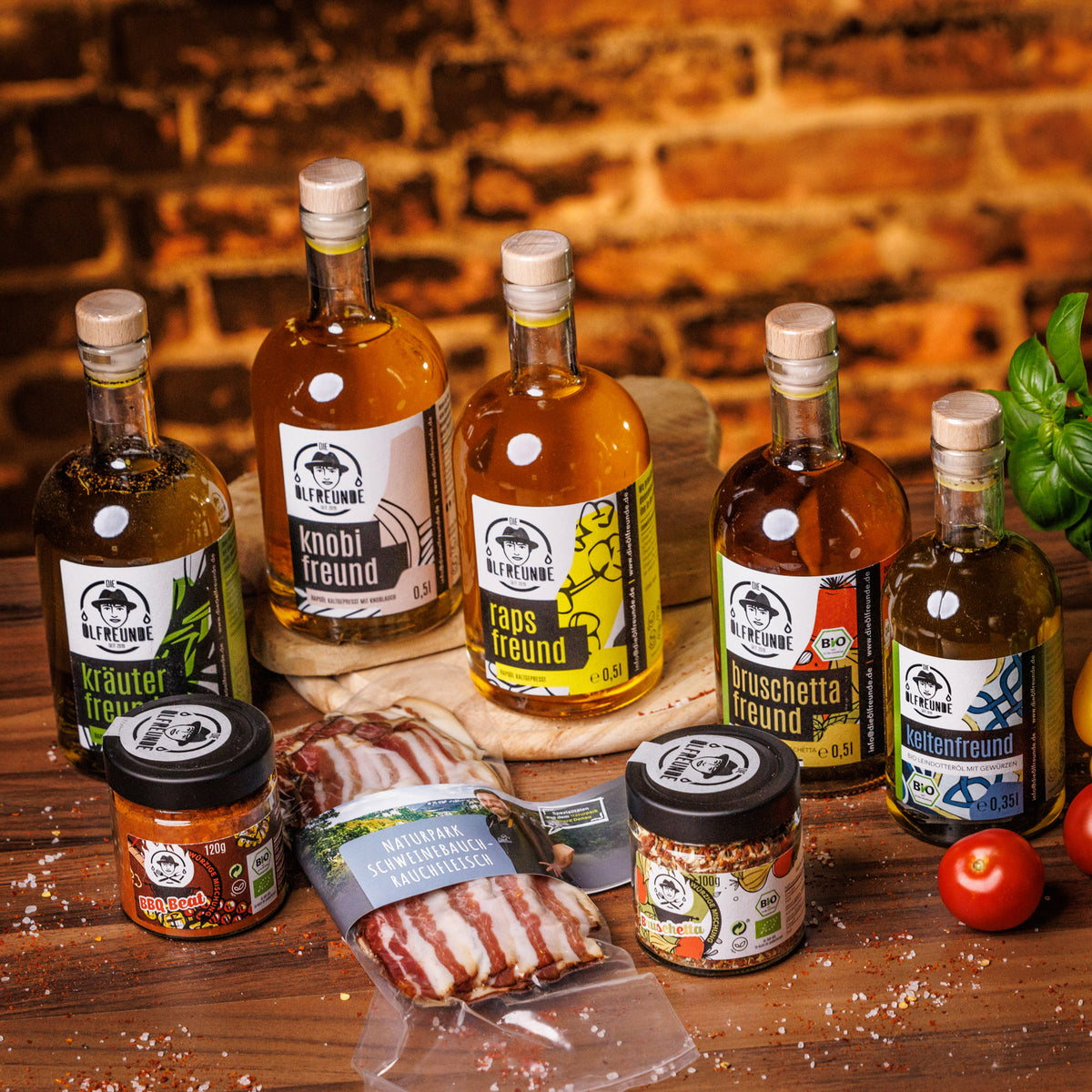 Eine Flasche Kräuteröl, Knoblauchöl, Rapsöl, Bruschetta-Öl, Leindotter-Öl mit Gewürzen, Ein Glas BBQ-Gewürzmischung und Burschetta-Gewürzmischung der Marke Die Ölfreunde und eine Packung Schweinebauch Rauchfleisch der Marke Naturpark Metzgerei Bosch, auf einem holzigen Untergrund, neben frischem Gemüse.